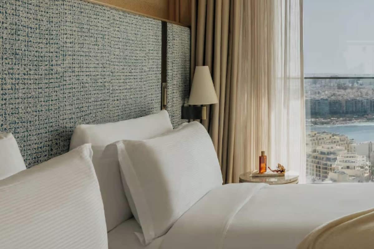 Quarto de hotel com uma cama grande, uma mesa de cabeceira, uma luminária e vista para a cidade através de uma janela ampla.