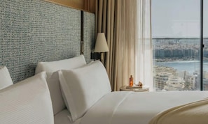 A hotel room with a large bed, a nightstand, a lamp, and a view of a cityscape through a large window.