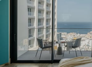 A balcony with a table and chairs, a bed, and a view of a cityscape and sea.