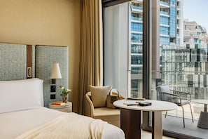 A hotel room with a large bed, a round table, a chair, a sofa, a lamp, a nightstand, and a view of the city through large windows.