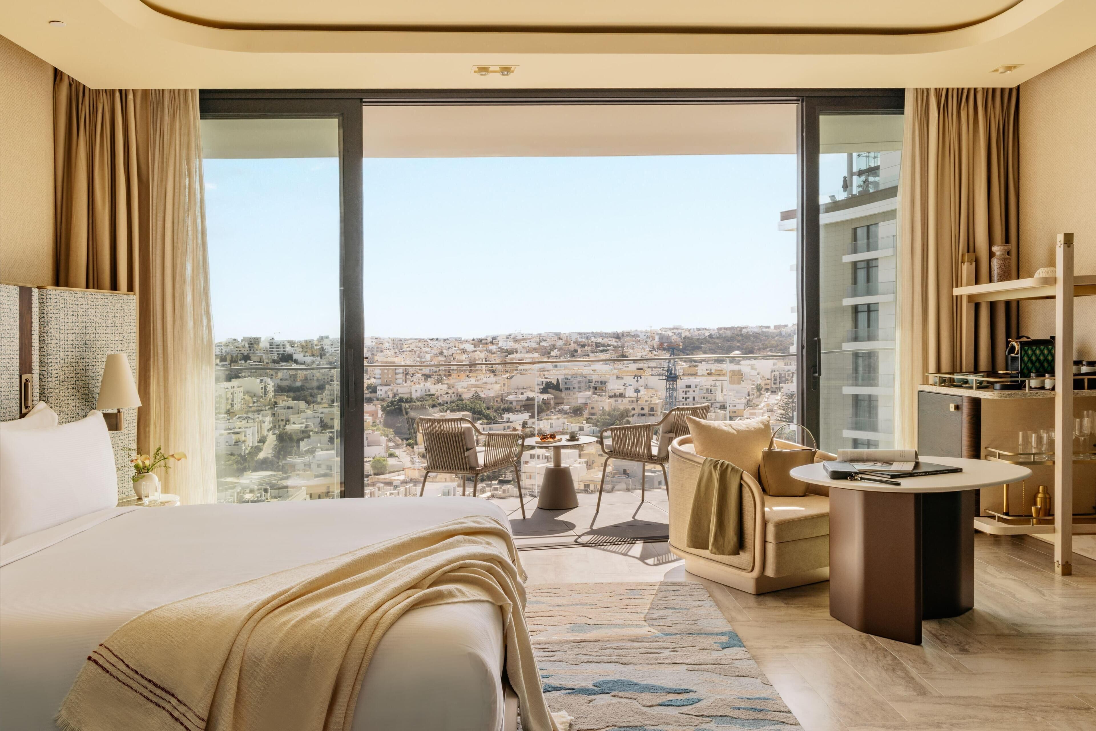 Quarto de hotel moderno com uma cama grande, um espaço de estar com uma mesa e vista para a cidade.