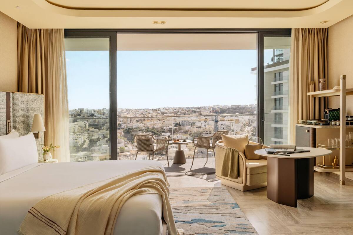 A modern hotel room with a large bed, a seating area with a table, and a view of the city.