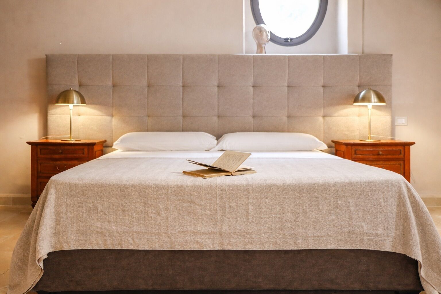 Un letto rifatto con precisione, con una testiera beige, due abat-jour e un libro sul letto.