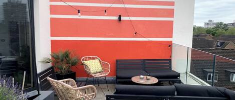 A balcony with a red and white wall, wicker chairs, black sofas, and a small table.