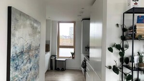 Uma cozinha pequena com um quadro abstrato grande à esquerda, uma janela com vista para edifícios e uma estante preta com plantas à direita.