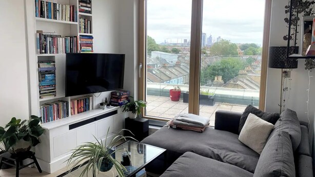 Uma sala de estar moderna com uma grande TV de tela plana, uma estante cheia de livros, um sofá cinza e vista para a cidade através de grandes janelas.