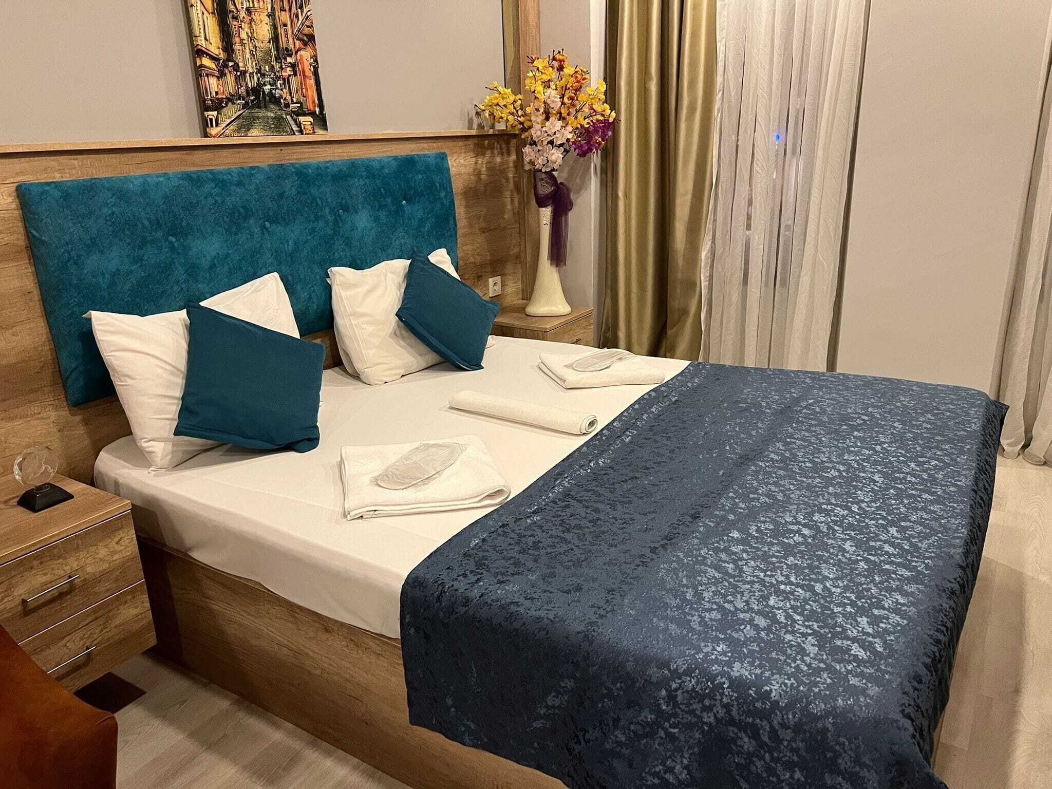 A neatly made bed with a blue headboard and pillows, a vase with flowers, and a painting on the wall.