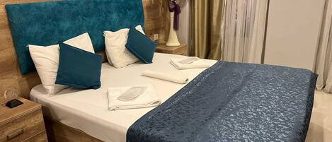 A neatly made bed with a blue headboard and pillows, a vase with flowers, and a painting on the wall.