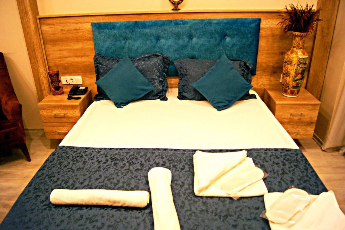 A neatly made bed with a blue headboard, two pillows, and a blanket. There is a wooden nightstand with a telephone and a decorative vase.