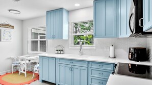 Une cuisine avec des meubles de couleur bleue, un plan de travail blanc, une machine à café et une fenêtre avec des stores.