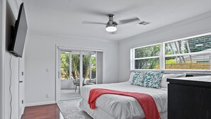 Une chambre avec un lit, un ventilateur de plafond, une fenêtre donnant sur un espace verdoyant et une porte menant à un espace extérieur.