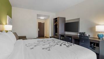A hotel room with a large bed, a desk with a flat-screen TV, a wardrobe, and a seating area with a chair.