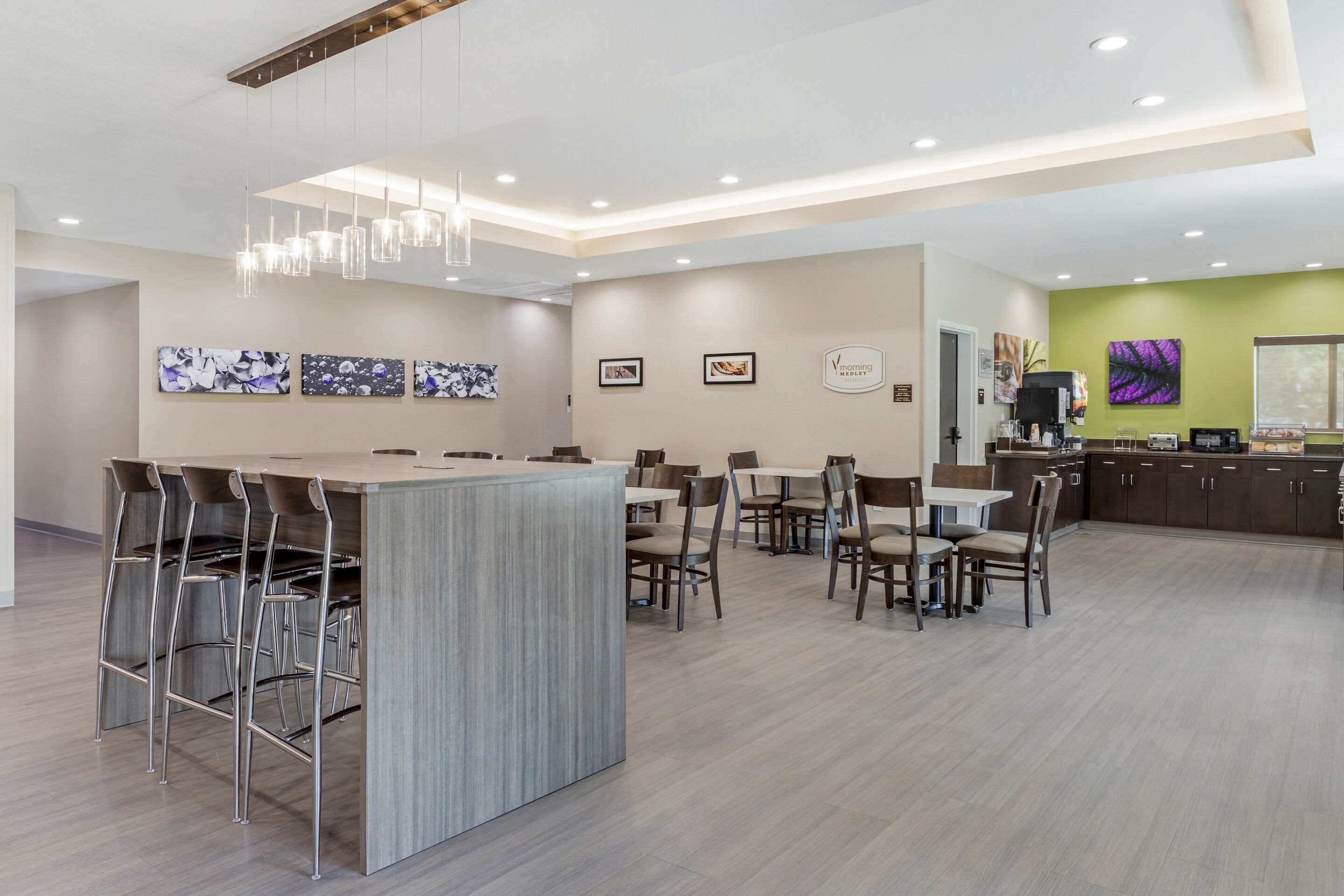 A modern hotel lobby with a bar area, dining tables, and a kitchen in the background.