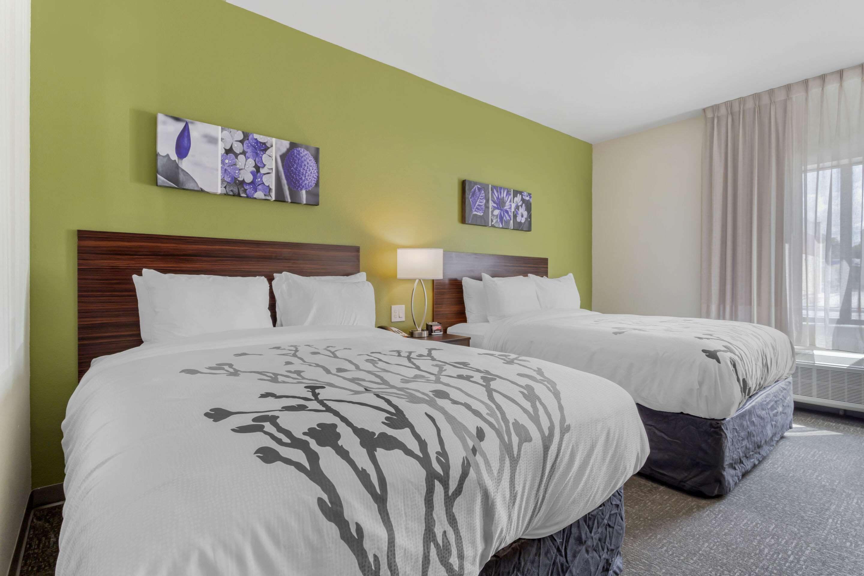 Quarto de hotel com duas camas, paredes verdes, obras de arte com motivos florais e uma janela grande com cortinas.
