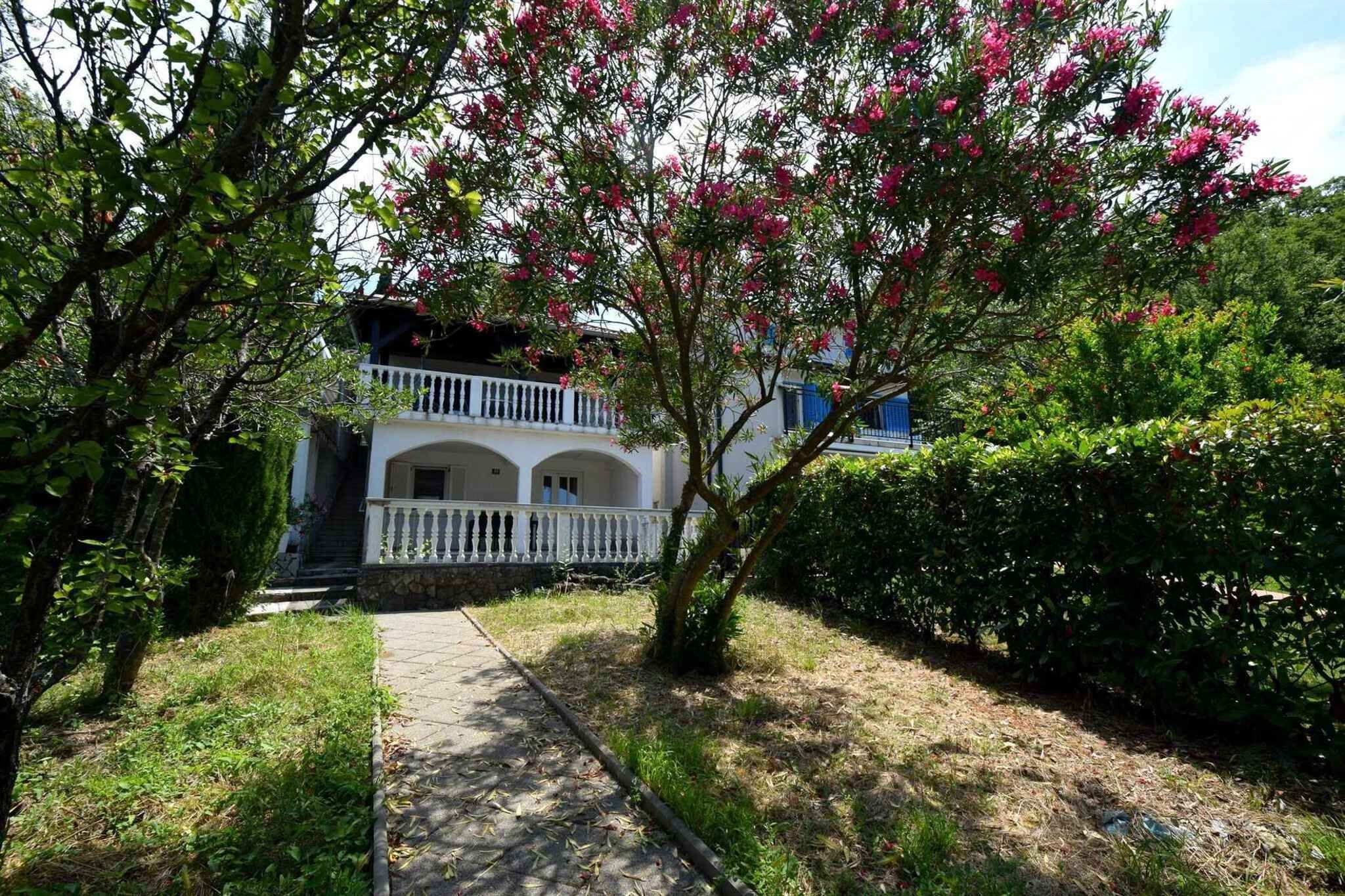 A two-story building with a white balcony, surrounded by greenery and a pathway.