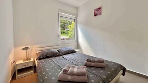A bedroom with a bed, a nightstand, a lamp, and a window with a view of greenery. - Holiday apartment mit Klimaanlage und Balkon (Jadranovo)