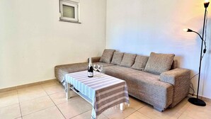 A living room with a sofa, a small table with a bottle and glasses, a floor lamp, and a window with blinds. - Holiday apartment modern and well equipped (Jadranovo)