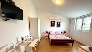 A bedroom with a bed, bedside tables, a desk with chairs, a TV, and a window with blinds. - Studio the sea with air conditioning (Jadranovo)