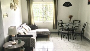 A small, well-lit room with a sofa, a round table with chairs, and a window with curtains. - Serenity 2-bed Apartment in Port Antonio, Portland (Port Antonio)