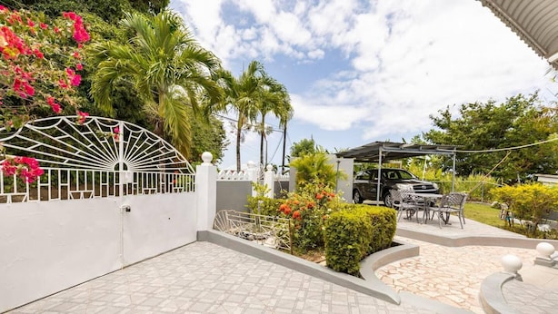 A gated outdoor area with a patio, a car parked under a covered area, and a white fence with a decorative gate. - Serenity 2-bed Apartment in Port Antonio, Portland (Port Antonio)