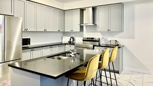 A modern kitchen with stainless steel appliances, a central island with bar stools, and white cabinetry. - 4-Bedroom House (Caledonia)