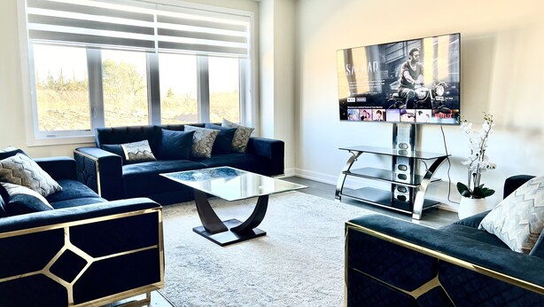 A modern living room with a dark blue sofa set, a glass coffee table, and a flat-screen TV mounted on the wall. - 4-Bedroom House (Caledonia)
