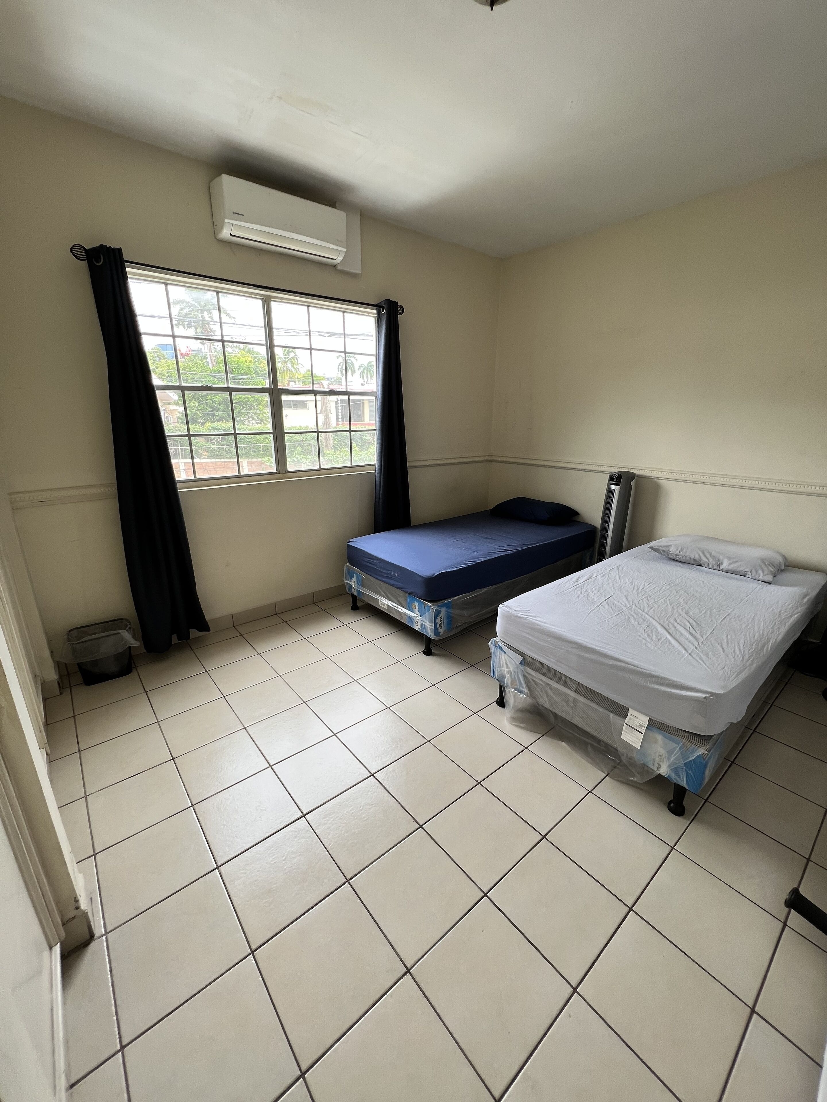 Une petite chambre propre, avec deux lits, une fenêtre avec des rideaux et un appareil de climatisation.