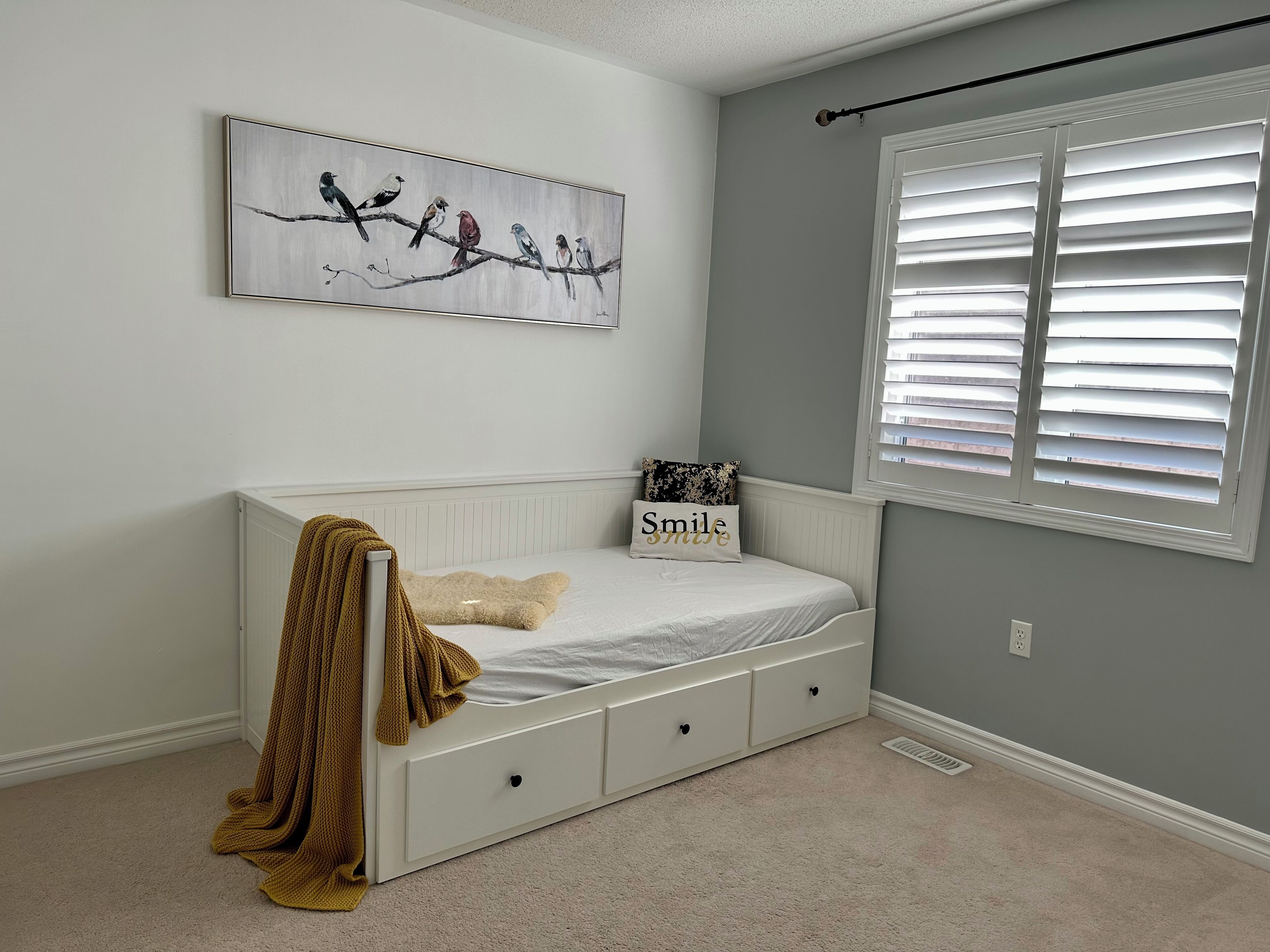 Une chambre avec un lit blanc, un tableau représentant des oiseaux sur le mur, une fenêtre avec des stores et un coussin orné du mot « Sourire ».