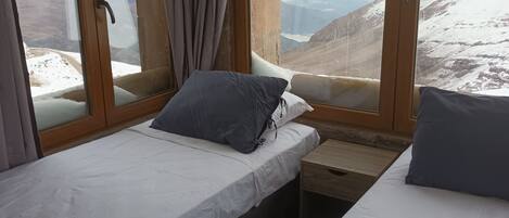 A room with two beds, a wooden nightstand, and large windows offering a mountain view.