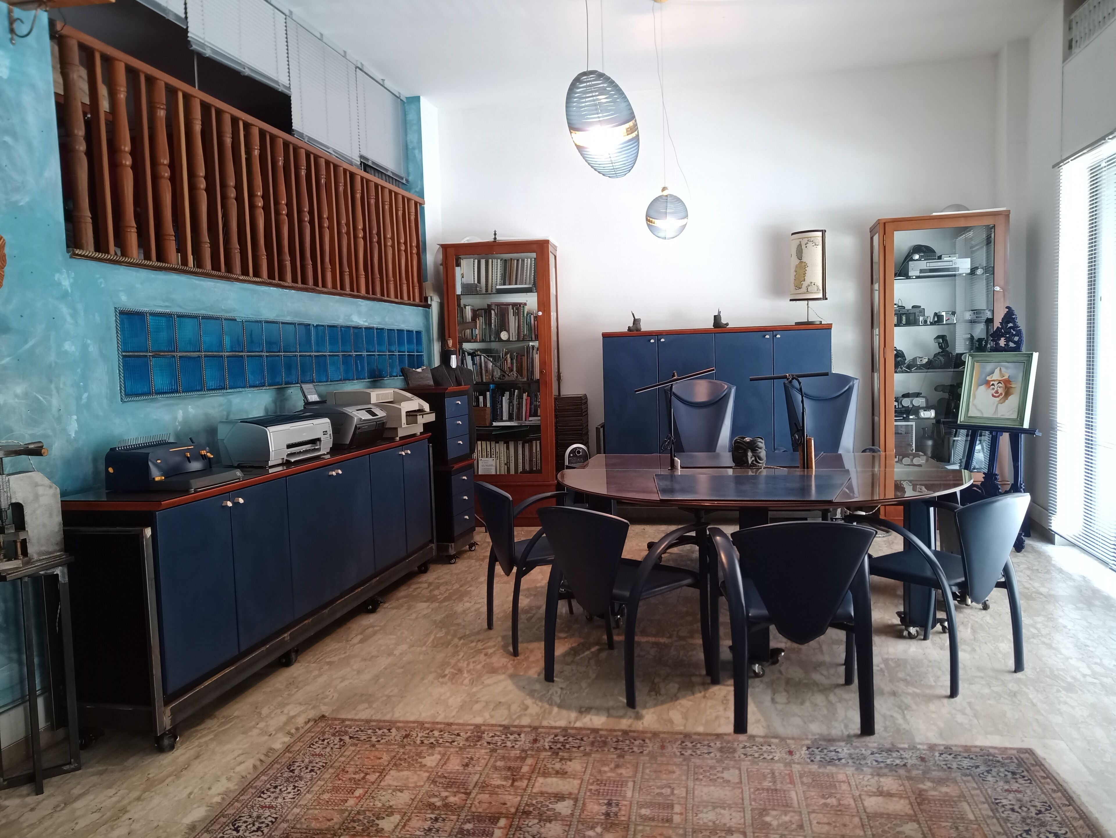 A room with a wooden desk, a bookshelf, a dining table with chairs, and a patterned rug.