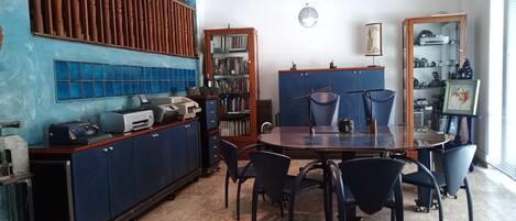 A room with a wooden desk, a bookshelf, a dining table with chairs, and a patterned rug.