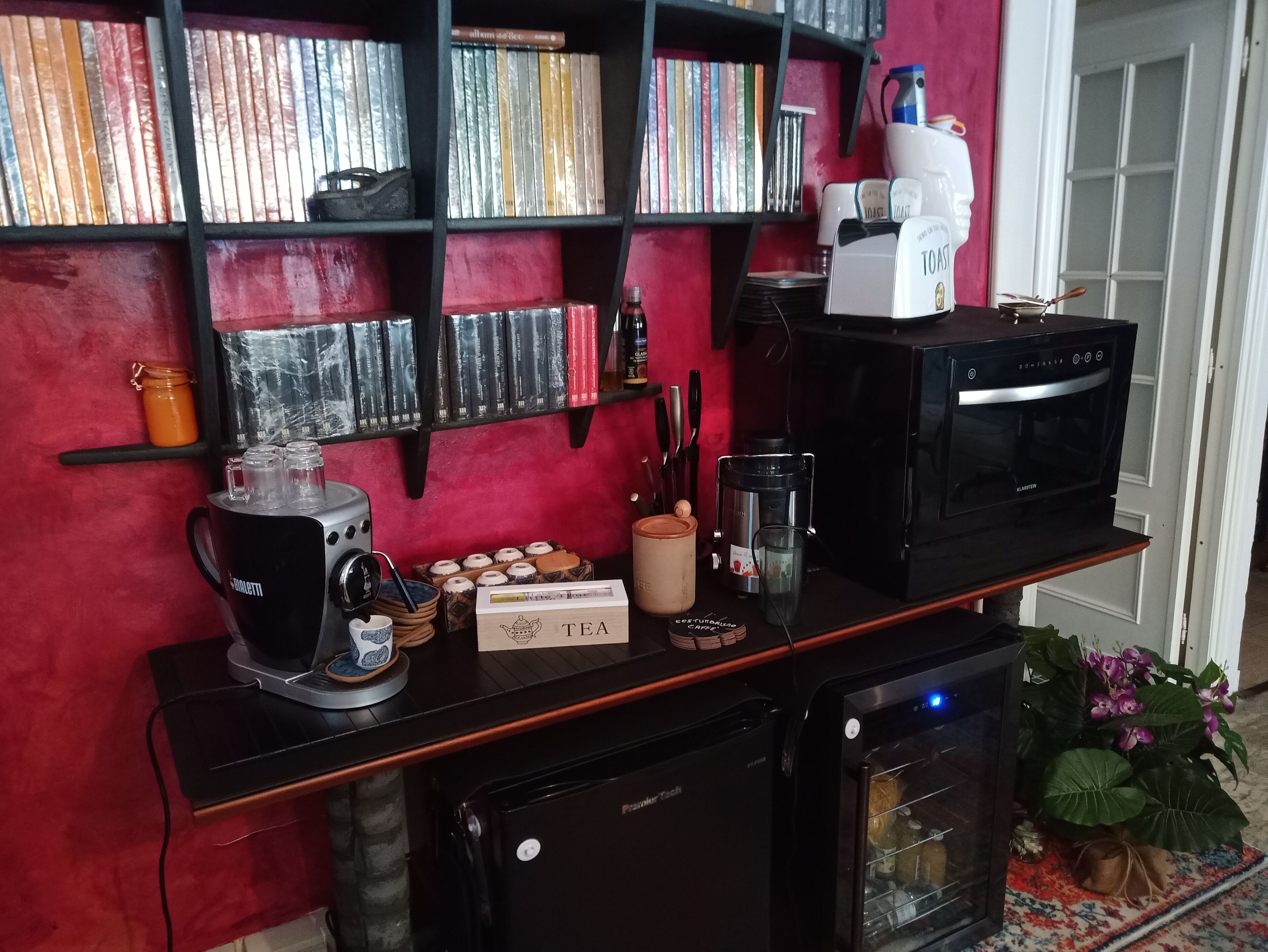 Un piano di lavoro in cucina con una macchina per caffè, un tostapane e un forno a microonde. Dietro al piano, si vedono dei libri su uno scaffale.