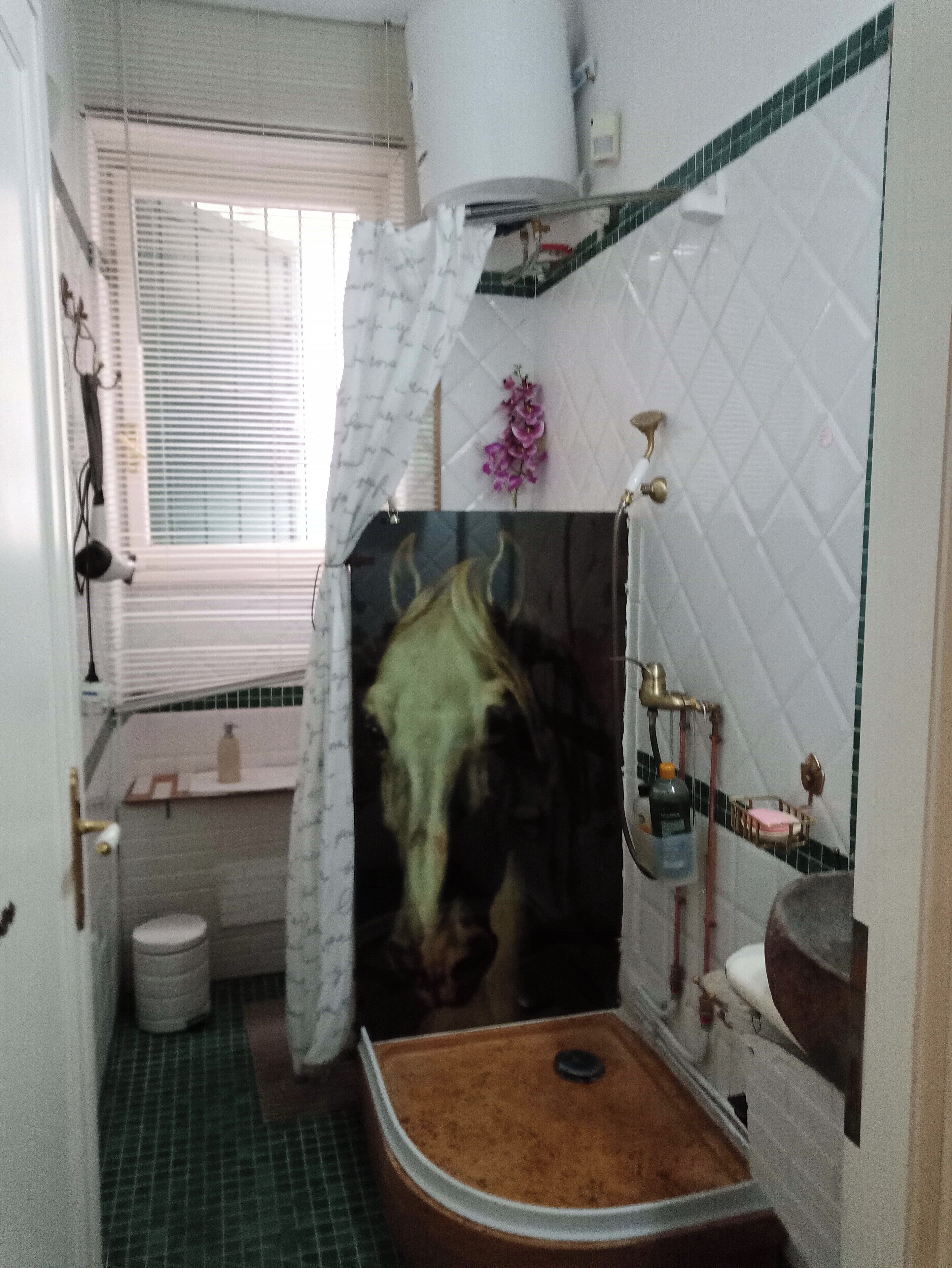 Une salle de bain avec douche, toilettes et lavabo. On y trouve une fenêtre avec stores, un porte-serviettes et une petite étagère contenant des articles de toilette.