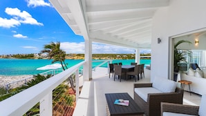 Un espace extérieur abrité, doté d’une piscine, d’une table à manger et de chaises, offrant une vue sur une mer turquoise et une plage.