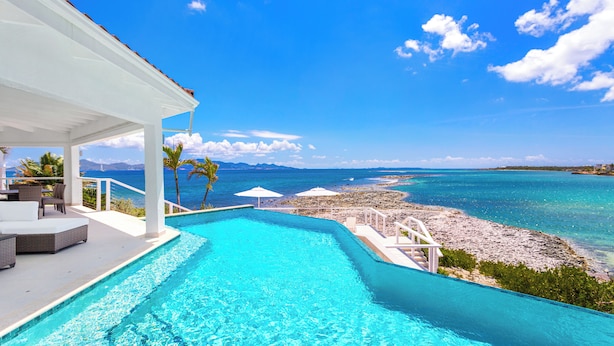 Une piscine avec vue sur l’océan et la plage, un espace de détente couvert avec des chaises et une table, et une structure blanche surmontée d’un toit rouge.