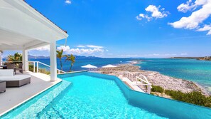 Ein Pool mit Blick auf den Ozean und den Strand, eine überdachte Sitzgelegenheit mit Stühlen und Tisch sowie eine weiße Struktur mit rotem Dach.