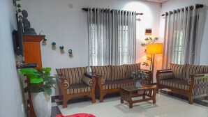 Sala de estar com dois sofás de madeira, uma mesa de centro de vidro e um pufe vermelho. Há cortinas, um abajur e uma estátua de Buda na parede.