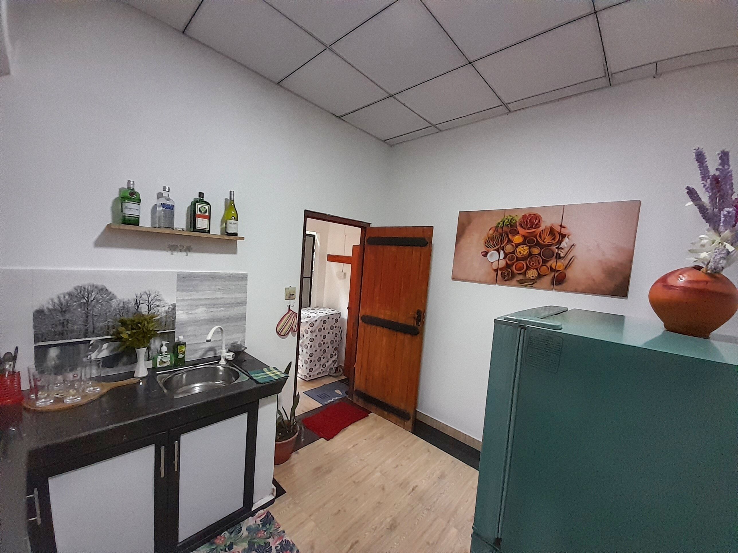 A kitchen with a sink, countertop, and a wooden door leading to another room. There are bottles on a shelf above the counter and a vase with flowers on a cabinet.
