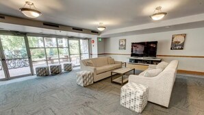 Eine geräumige Lobby mit einem beigefarbenen Sofa, einem Couchtisch und gemusterten Hocker. Ein Fernseher ist an der Wand montiert, und große Fenster bieten Blick auf Fahrräder draußen.