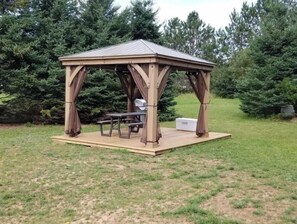 Relax in a serene outdoor setting surrounded by trees and shrubs, with a picnic table and benches under a gabled roof.