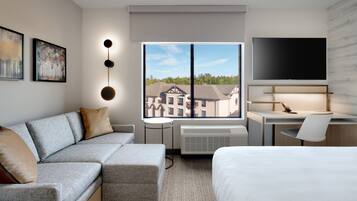 Una habitación de hotel moderna con un sofá gris, una cama, un escritorio con una silla, un televisor de pantalla plana y un ventanal que ofrece vistas a edificios y árboles.