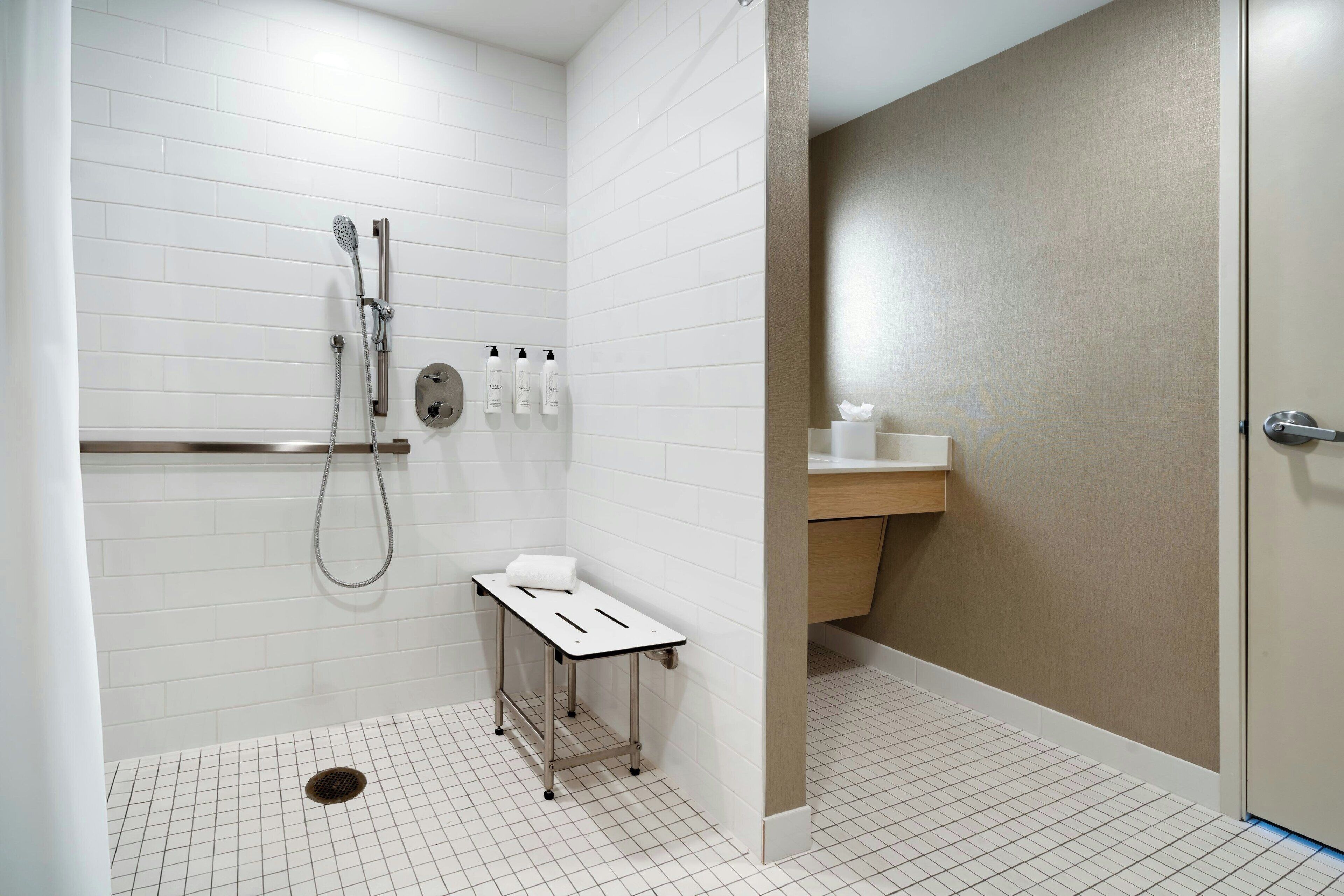 Un baño moderno con paredes de azulejos blancos, ducha con cabezal de ducha manual, una mesita con una toalla y un mostrador de madera con lavamanos.
