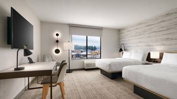Habitación de hotel con cama, escritorio, silla y un amplio ventanal que ofrece vistas a edificios y árboles.