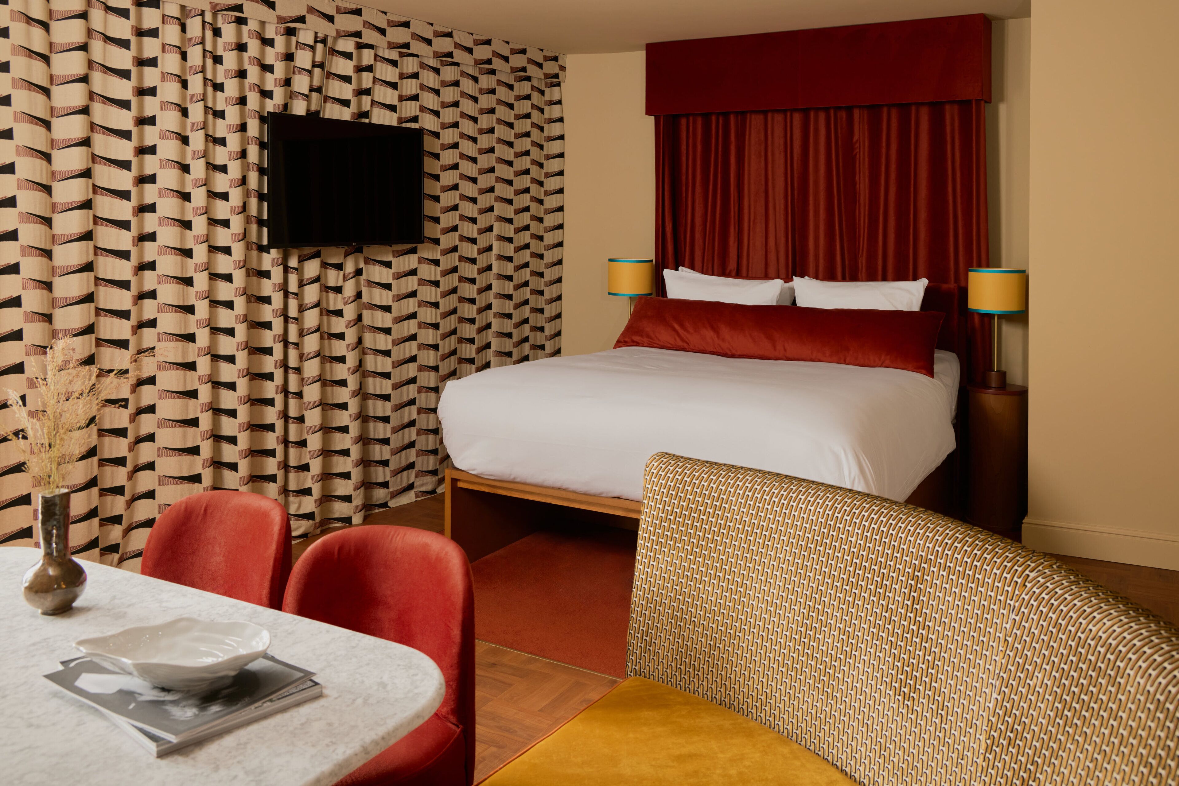 A hotel room with a bed, a television mounted on the wall, a dining table with chairs, and a vase with dried plants.