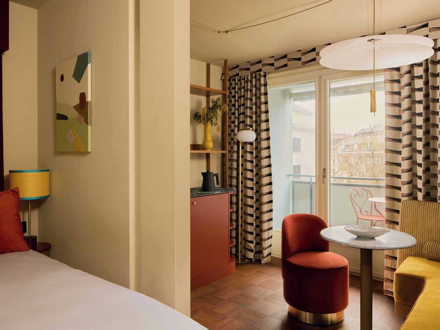 Quarto de hotel com cama, uma pequena mesa, uma cadeira vermelha, um sofá amarelo, cozinha e varanda com cortinas estampadas.