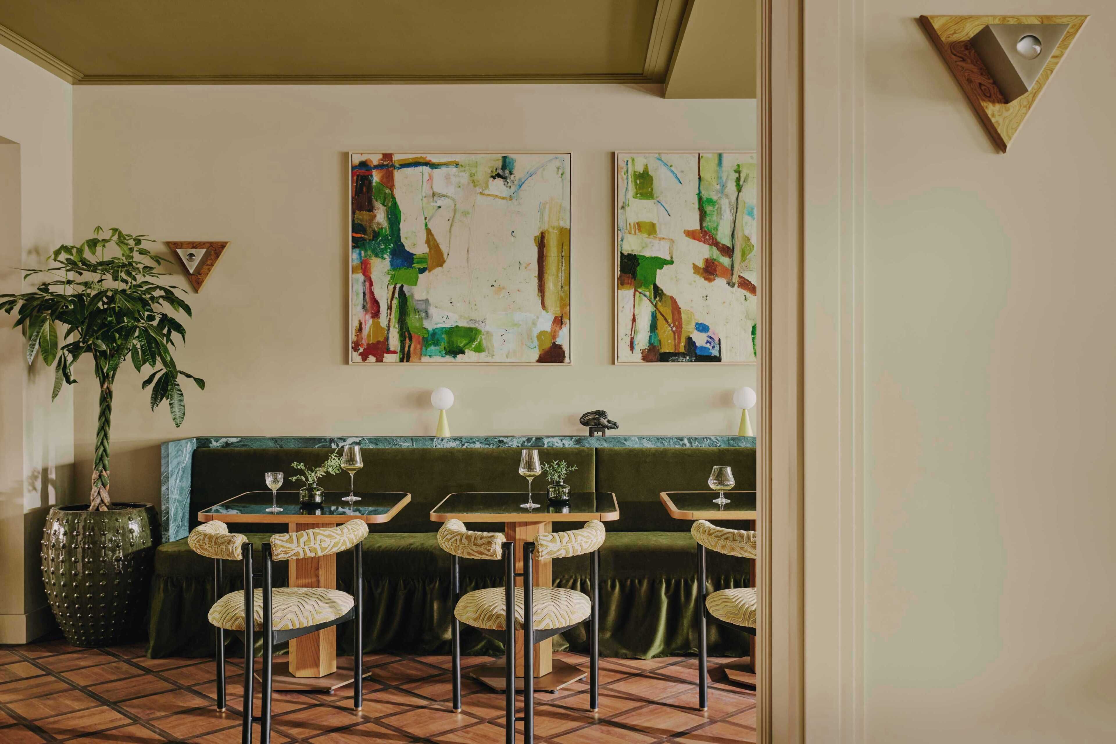 L’intérieur d’un restaurant présente un banc vert rembourré, trois tables rondes avec des chaises et des peintures abstraites accrochées au mur.