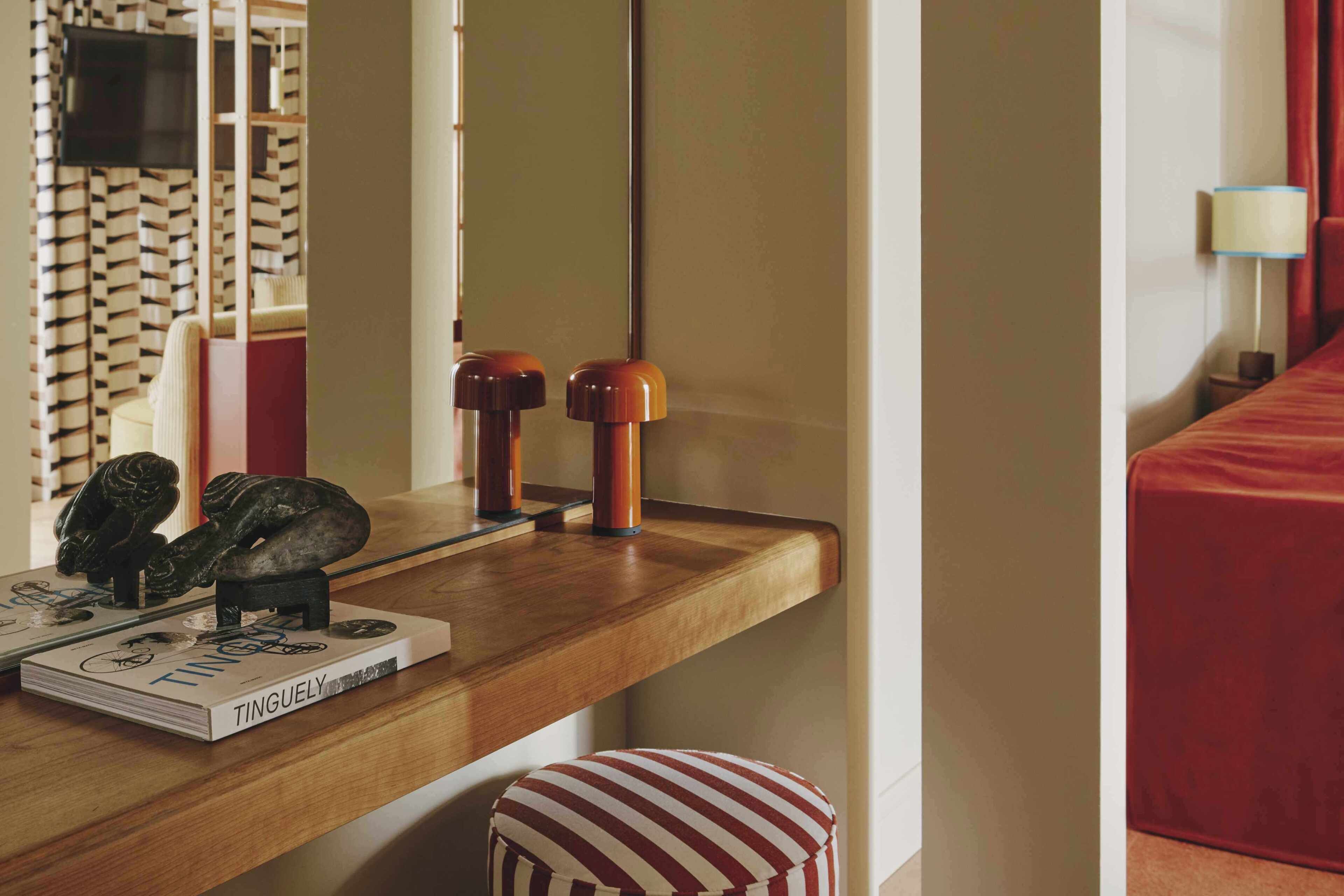 A hotel room with a wooden desk, a mirror, a red-striped ottoman, and a television on the wall.