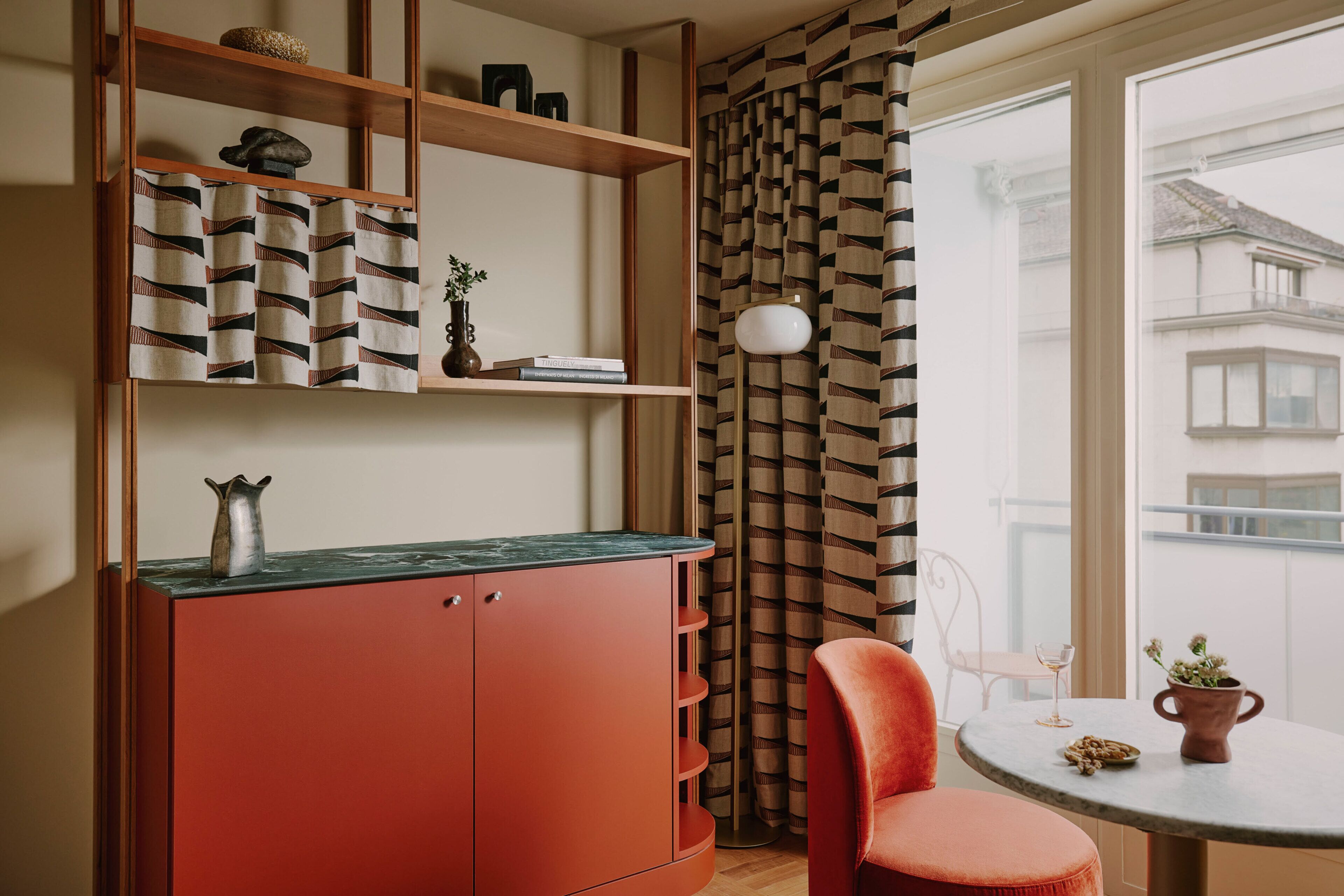 A room with a red cabinet, shelves with books, a chair, a table with a vase, and a view of buildings through a window.