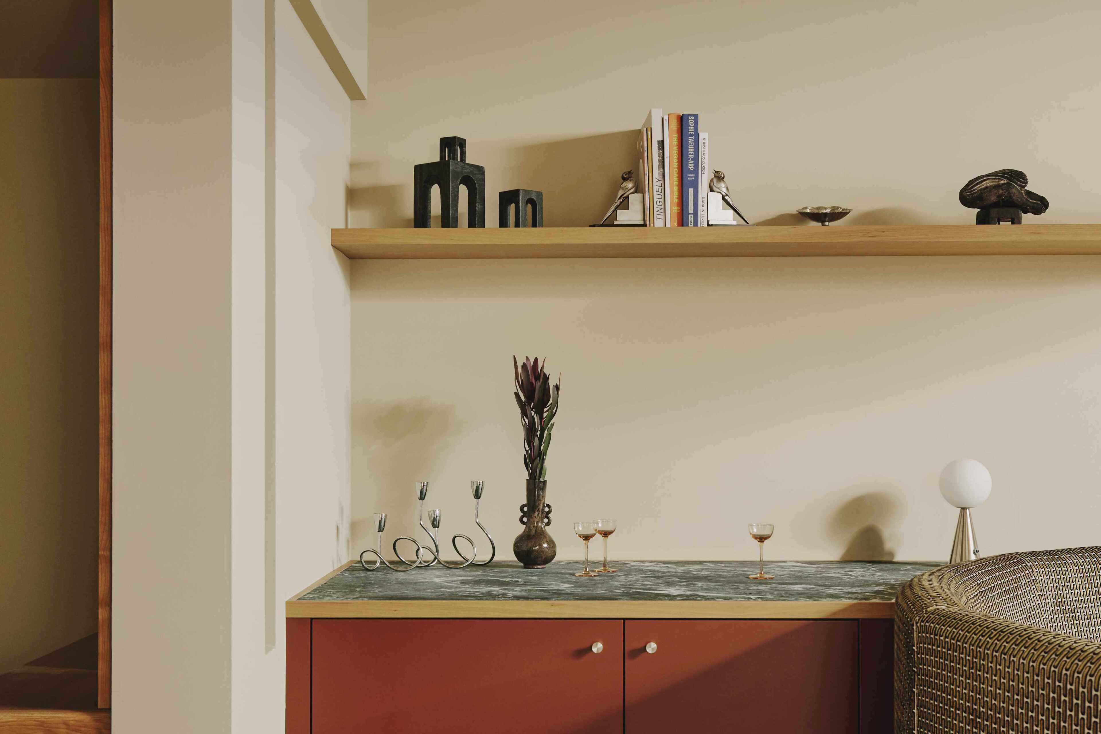 Una habitación con un estante de madera, un jarrón con plantas secas, una mesita con un vaso y una silla de mimbre.
