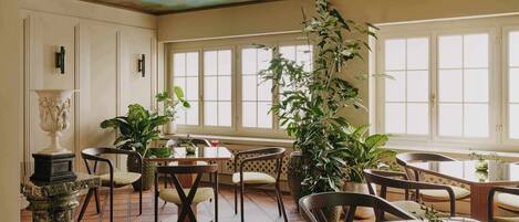 A dining area with multiple tables and chairs, large windows, and potted plants.